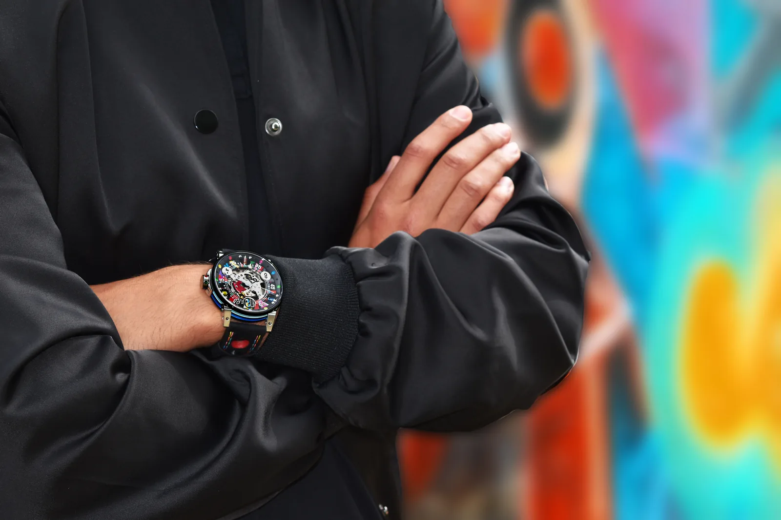 Bras croisés en blouson noir, montre Art Car squelette au poignet, fresque colorée en fond